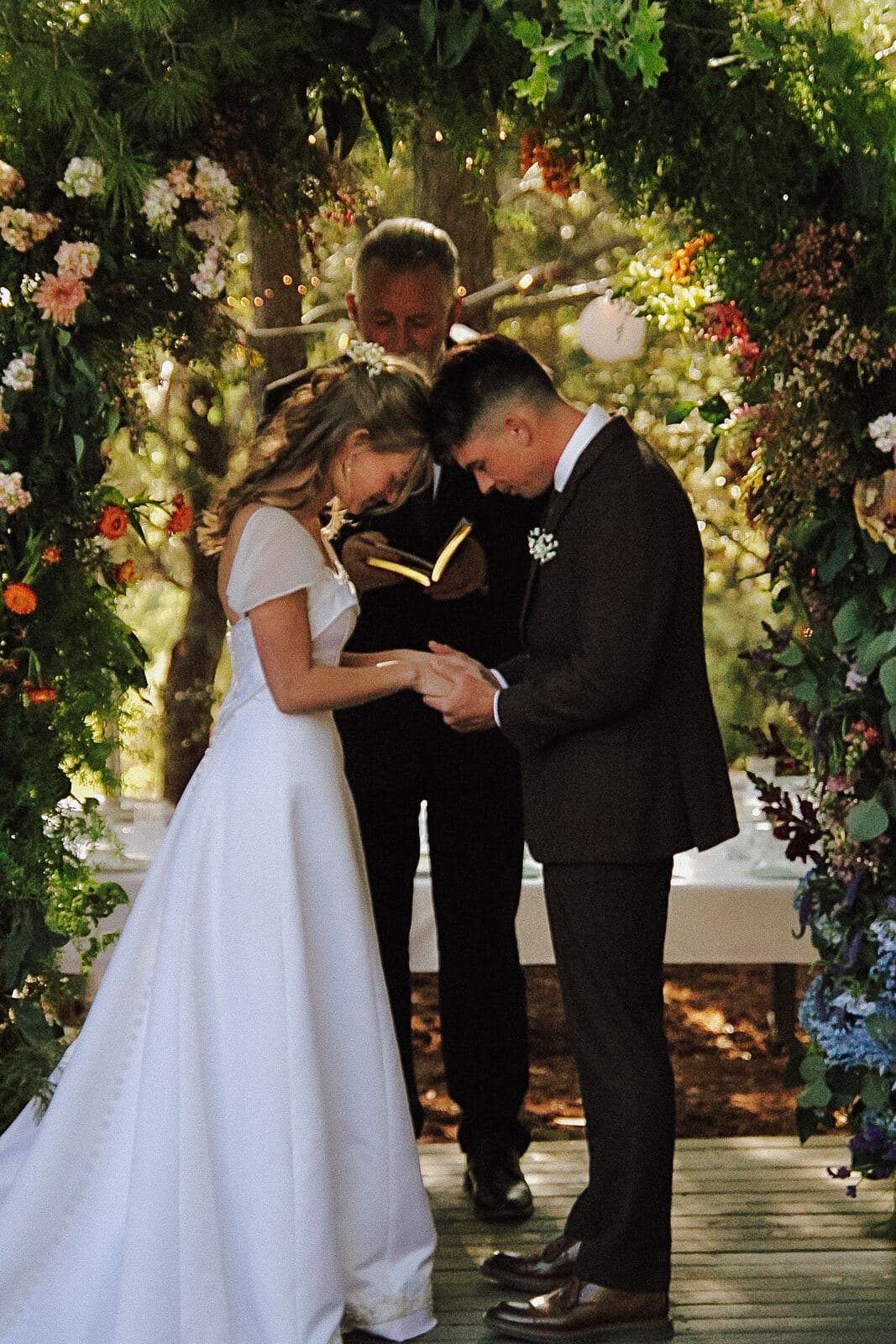bride and groom bowing their heads in prayer beneath a lush floral ceremony arch