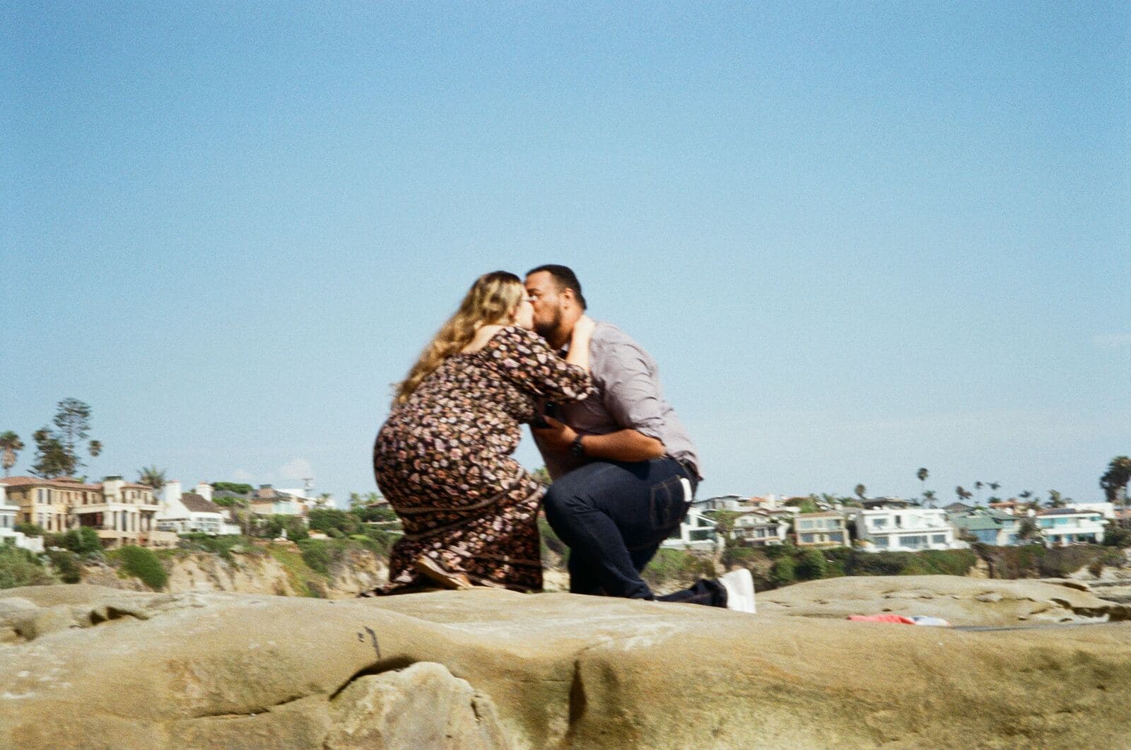 newly engaged couple kissing while kneeling on sandstone rocks with a coastal town behind them