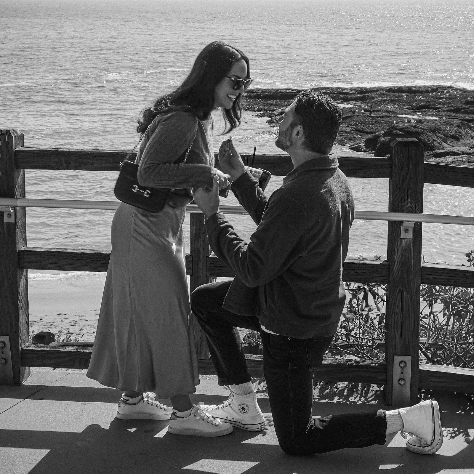 black and white photo of a man on one knee proposing to a smiling woman at a beachside overlook