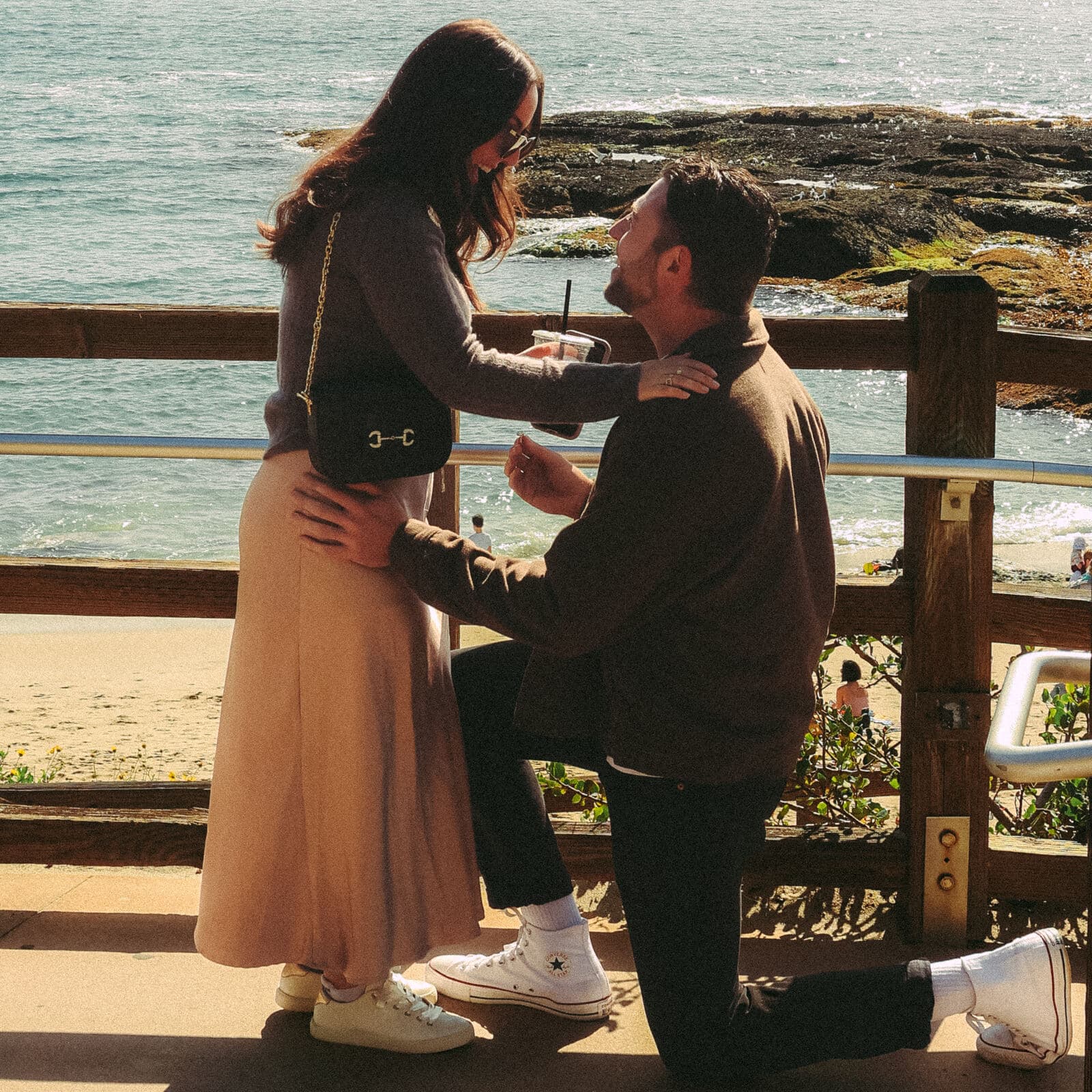 man kneeling with a ring as a woman holding an iced coffee smiles down at him by the ocean