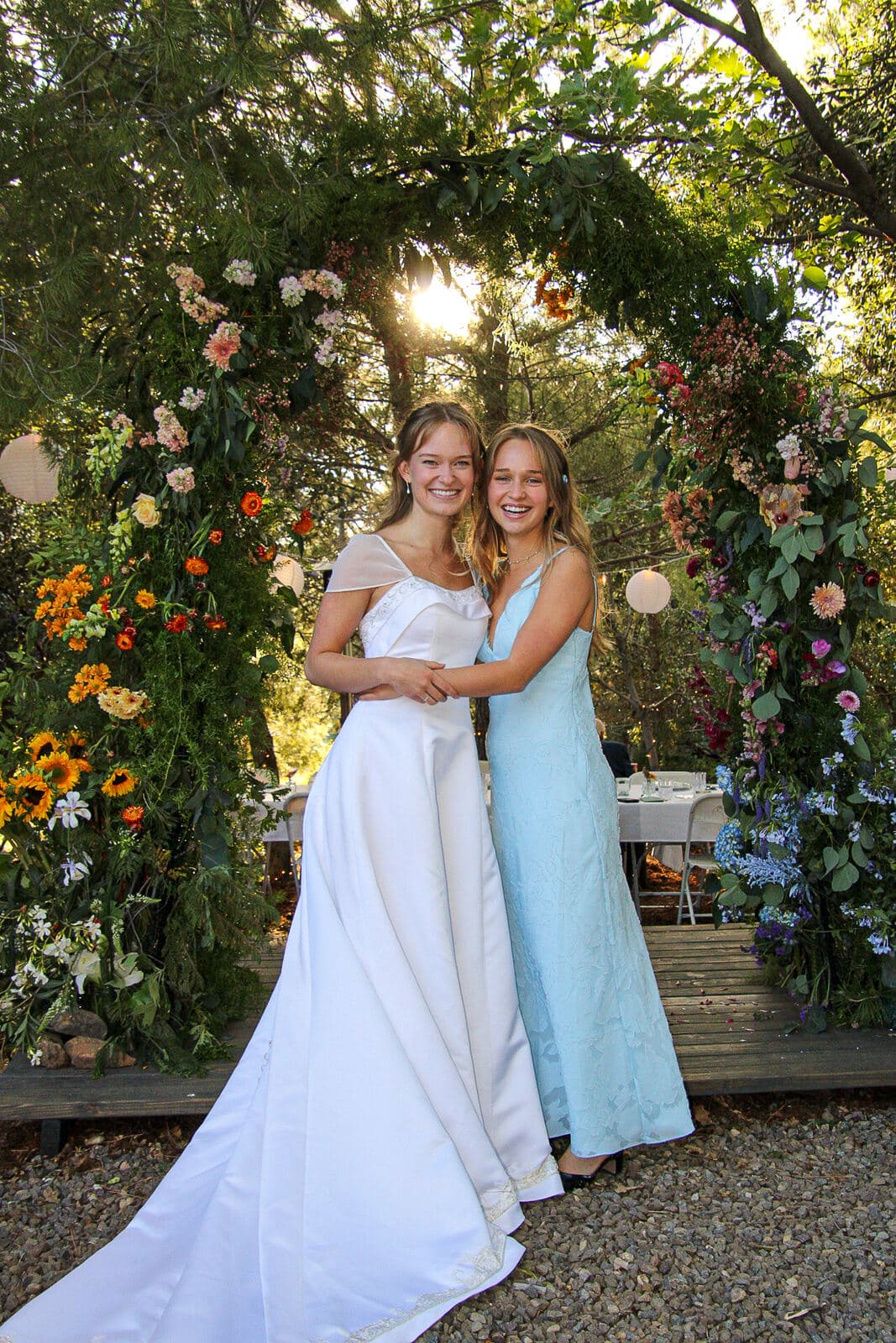 bride hugged from behind by a bridesmaid in blue in front of a sunlit floral arch