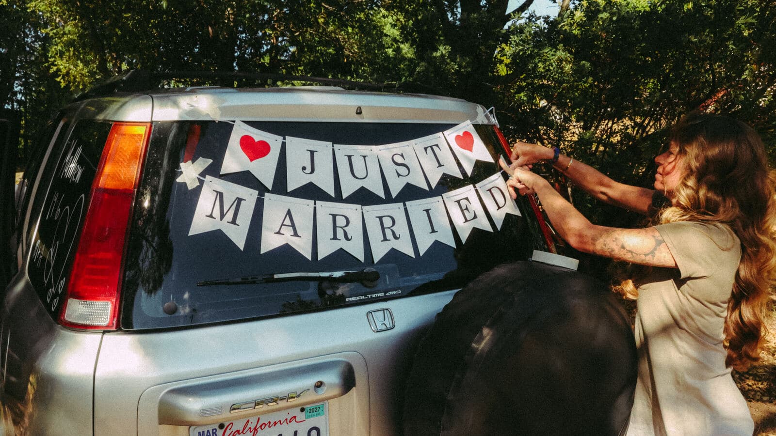 woman hanging a Just Married banner across the rear window of a getaway car under the trees